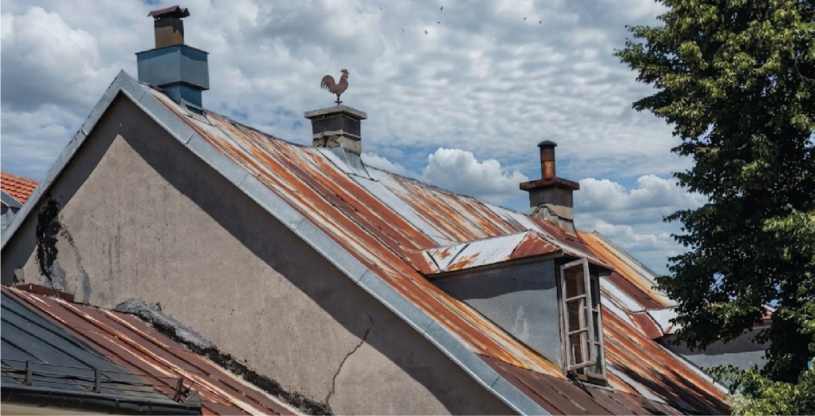 Old metal roofs streaked with rust beneath a cloudy sky, chimneys and a weather-vane cockerel along the ridge